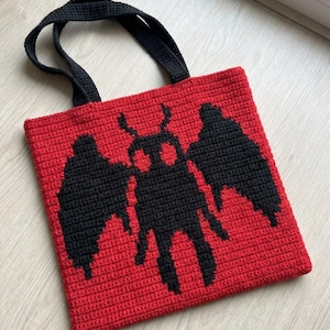 Puede incluir: Bolso tote rojo tejido a ganchillo con asas negras y un diseño de polilla negra. El bolso es de forma cuadrada y está hecho a mano. Ideal para llevar libros, comestibles u otros artículos.