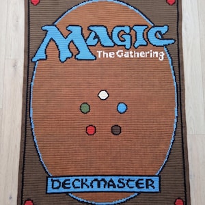 May include: A rectangular crochet blanket featuring the Magic The Gathering card back design. The brown background displays the blue Magic logo, the text The Gathering, and the word DECKMASTER. Five colored circles are centered in the middle.