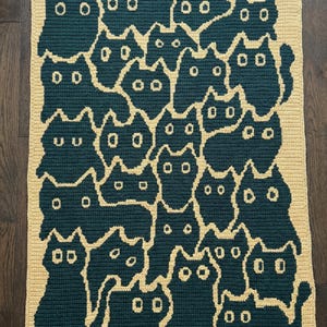May include: A crocheted blanket with a repeating pattern of dark teal cat shapes on a light yellow background. The blanket is rectangular and looks to be made from soft yarn. A cosy home decor item, measuring approximately 120cm x 150cm.