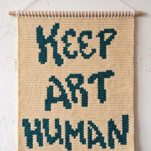 May include: A rectangular crochet wall hanging in cream yarn featuring the dark teal text KEEP ART HUMAN. The piece is suspended from a light wooden dowel with a white cord, showcasing a textured stitch pattern throughout the entire design.