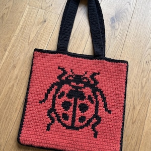 Könnte beinhalten: Eine quadratische Häkeltasche mit rotem Körper und einer schwarzen Marienkäfer-Grafik in der Mitte. Die Tasche hat zwei schwarze Häkelgriffe und einen schwarzen Randbesatz an den Kanten, gefertigt mit einem sichtbaren Garnstichmuster.