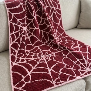 May include: A rectangular crochet blanket featuring a repeating spider web pattern in deep burgundy and light pink. The textile has a textured surface with a consistent stitch throughout and is finished with a light pink border along the edges.