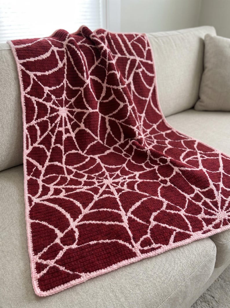 May include: A rectangular crochet blanket featuring a repeating spider web pattern in deep burgundy and light pink. The textile has a textured surface with a consistent stitch throughout and is finished with a light pink border along the edges.