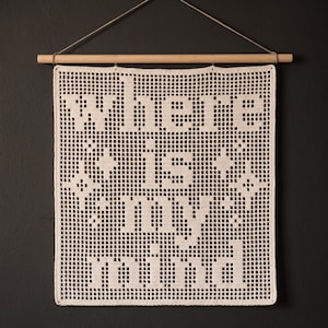 May include: A cream-colored crochet wall hanging featuring a grid pattern with the text "Where is my mind" and four-pointed star shapes. The textile is suspended from a horizontal wooden dowel with a thin string attached for hanging purposes.