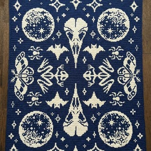 May include: A rectangular crochet blanket featuring a dark blue background with a cream-coloured pattern. The design includes two bird skulls, four moons, four moths, four bats, four spiders, and various star shapes arranged in a symmetrical layout.