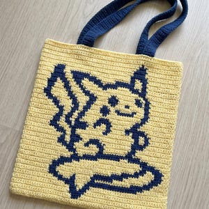 Könnte beinhalten: Eine handgefertigte Häkel-Tragetasche in blassem Gelb mit einem dunkelblauen Grafikdesign einer Zeichentrickfigur mit spitzen Ohren und einem blitzförmigen Schwanz. Die Tasche verfügt über zwei dunkelblaue Häkelgriffe an der Oberkante.