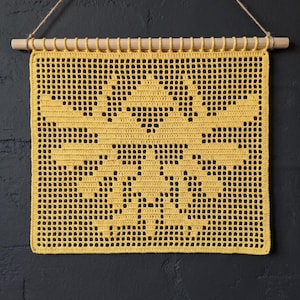 Op de afbeelding: Een vierkante gehaakte wandhanger in felgeel met een gedetailleerd bijenmotief, gemaakt met een rasterachtig opengewerkt patroon. Het textiel hangt aan een lichtgekleurde houten stok met een dun touwtje eraan bevestigd om op te hangen.