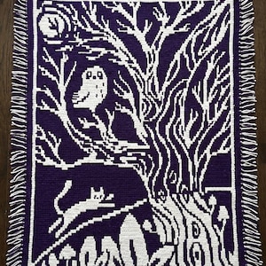 Puede incluir: Una manta rectangular de ganchillo con un patrón de mosaico superpuesto en color morado oscuro y blanco. El diseño muestra un árbol grande, un búho posado en una rama, un gato, setas y una luna. La manta tiene flecos blancos y morados.