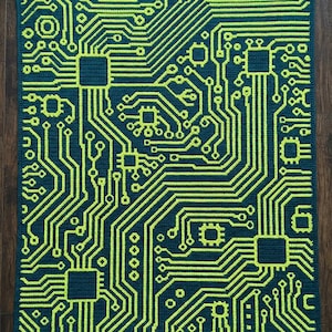 May include: A crocheted blanket featuring a circuit board design. The blanket has a dark teal background with a bright neon green circuit pattern. The design includes squares and lines, resembling the internal components of a computer.