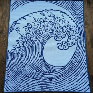 May include: A crocheted blanket featuring a navy blue and light blue design of a large ocean wave. The wave is depicted with intricate details, including swirling lines and frothy crests. The blanket is rectangular and appears to be handmade.