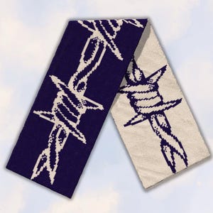 May include: A rectangular knit scarf featuring a repeating pixelated barbed wire pattern. One side displays a dark navy blue background with a white barbed wire design, while the other side reverses the color scheme with a white background and navy blue.