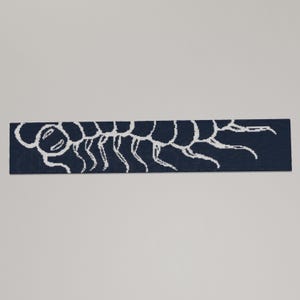 May include: A navy blue rectangular rug featuring a white centipede design. The centipede is depicted in a simple, stylized manner, with a series of connected body segments and numerous legs. The rug is likely made of a soft material.