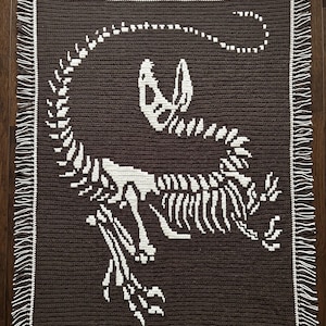 May include: A rectangular crochet blanket featuring a dark brown background with a large, white, detailed dinosaur skeleton motif centered on the fabric. The blanket is finished with a white border and decorative fringe along the two shorter sides.