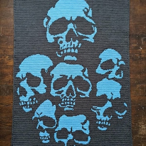 Op de afbeelding: Een donkergrijze gehaakte deken met een patroon van blauwe schedels. De schedels zijn van verschillende groottes en zijn over de deken verdeeld. De deken is rechthoekig en lijkt handgemaakt te zijn, met een getextureerd oppervlak.