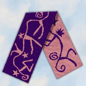 May include: A rectangular knit scarf featuring a reversible two-tone design. One side displays a deep purple background with light pink stick-figure patterns and star shapes, while the other side shows a light pink background with purple patterns.
