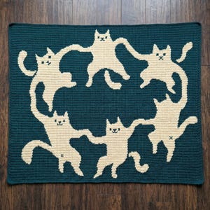 May include: A teal crocheted blanket featuring a repeating pattern of cream-colored cats in a circle. The cats have outstretched arms and legs, with simple facial features. The blanket is rectangular and appears handmade.