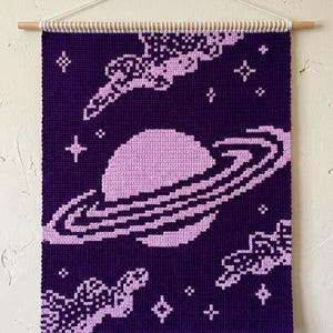 May include: A handmade crochet wall hanging depicting a space scene in purple and lavender. The design features a planet with rings, stars, and cloud formations. It is suspended from a wooden dowel with a white cord.