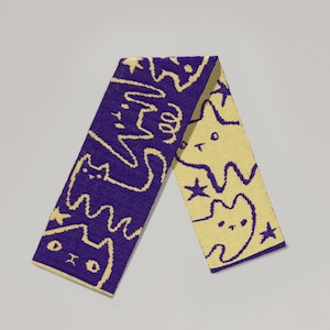 May include: A purple and yellow scarf with a repeating pattern of cartoon cats, stars, and abstract shapes. The scarf is folded in half, displaying the design on both sides. The scarf is made of a soft, knitted material.
