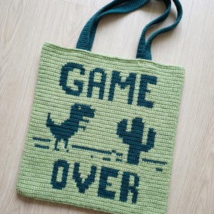 Może przedstawiać: Szydełkowa torba w kolorze jasnozielonym z ciemnoniebieskimi akcentami. Torba zawiera napis "GAME OVER" w dużych literach, a także dinozaura i kaktusa. Uchwyty są w kolorze ciemnoniebieskim.