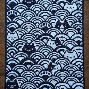 Può includere: Una coperta rettangolare all'uncinetto con un motivo ripetitivo di onde semicircolari blu navy scuro e azzurro. Il design incorpora volti di gatto stilizzati integrati nel motivo a onde, alternando tra colori scuri e chiari.