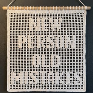 May include: A white crocheted wall hanging featuring a grid pattern with the text "NEW PERSON OLD MISTAKES" woven into the design. The piece is suspended from a light-colored wooden dowel with a white string attached for hanging on a wall.