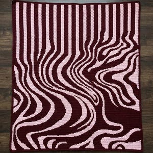 May include: A handmade crochet blanket with a unique design. The blanket features a pattern of vertical stripes that transition into a wavy, distorted design. The colors are a deep burgundy and a light pink, creating a striking visual effect.