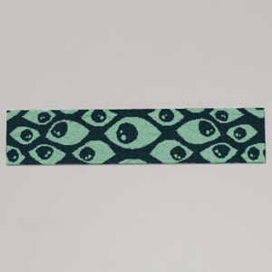 May include: A rectangular, knitted headband featuring a repeating pattern of stylized eyes. The eyes are a light teal color against a dark teal background. The headband is likely made of soft yarn or fabric.