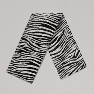 May include: A black and white zebra print scarf. The scarf is folded in a V-shape, showcasing the bold pattern. The fabric appears soft and textured, with the zebra stripes running across the length of the scarf.