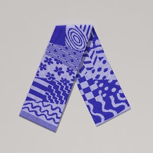 Puede incluir: Una toalla estampada azul y blanca con varios diseños geométricos, incluyendo flores, zigzags y círculos. La toalla está doblada por la mitad, mostrando los diferentes patrones en cada lado. La toalla está hecha de un material suave y absorbente.
