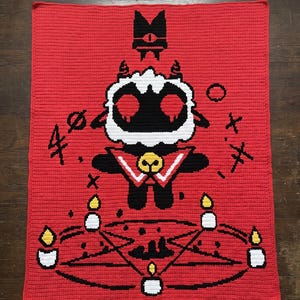May include: A rectangular crochet blanket featuring a red background with a central black and white lamb character wearing a bell. Below the character is a black pentagram surrounded by five white candles with yellow flames. Black symbols flank the lamb.