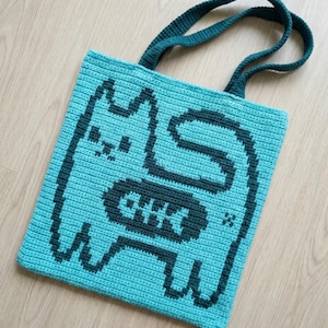 May include: A turquoise crochet tote bag featuring a pixelated cat design with a fish bone motif. The cat and fish bones are dark teal. The bag has teal handles and is made of a textured yarn.