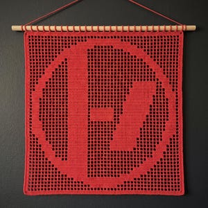 May include: A square red crochet wall hanging featuring a large letter P inside a circular border. The piece is constructed with a grid-like filet crochet pattern and is suspended from a light-colored wooden dowel with a thin red string.