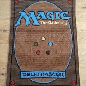 Puede incluir: Una manta rectangular de ganchillo con el diseño del reverso de una carta de Magic The Gathering. El fondo marrón muestra el logotipo azul de Magic The Gathering, el texto Deckmaster y cinco pequeños círculos de colores amarillo, azul, verde, rojo y marrón.