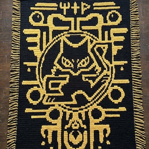 May include: A rectangular black and gold crochet blanket featuring an intricate overlay mosaic design of a central cat figure surrounded by geometric patterns and symbols. The blanket has a fringed edge with alternating black and gold yarn strands.