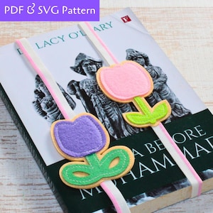 May include: Two felt flower bookmarks, one purple and one pink, with green leaves and stems. The bookmarks are attached to white and pink ribbon and are placed on a book.