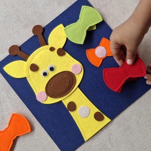 May include: A felt giraffe puzzle with a yellow head, brown spots, and a blue background. The puzzle includes three colorful bow ties in green, orange, and red. A hand is reaching for a bow tie.