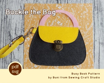 Può includere: Un modello di libro occupato in feltro con un design a forma di borsa. La borsa è principalmente grigio scuro con una patta gialla e un manico rosa. Il testo "Buckle the Bag" è visualizzato sopra la borsa. Un cinturino da polso giallo è attaccato alla borsa.
