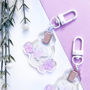 May include: Two clear acrylic keychains shaped like bottles with a white bunny inside and pink flowers. The bottles have a brown cork top and a silver metal ring.