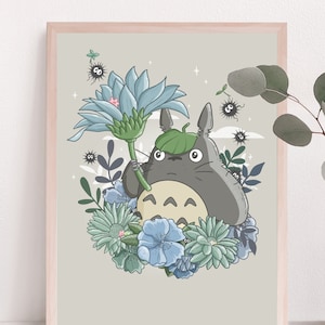 Op de afbeelding: Een ingelijst afdruk van een cartoonfiguur, Totoro, die een blauwe bloem vasthoudt. De achtergrond is lichtgrijs met een paar kleine witte wolken en zwarte en witte stippen. De afdruk is omgeven door een lichtbruine lijst.