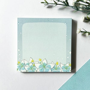 May include: A light blue sticky note with a white border and a floral design at the bottom. The flowers are white with blue centres and yellow centres. The background is a light blue with white glitter.