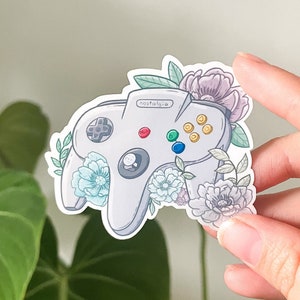 Könnte beinhalten: Ein weißer Nintendo 64-Controller mit floralen Akzenten. Der Controller hat einen hellblauen, roten, grünen und gelben Knopf. Der Text "Nostalgie" ist auf den Controller gedruckt.