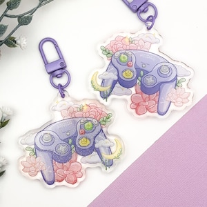 May include: Two purple GameCube controller keychains with floral and cloud designs. The keychains are made of clear acrylic and have a purple metal clasp.