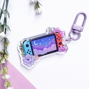 Puede incluir: Un llavero de acrílico transparente con una ilustración estilizada de una consola Nintendo Switch con un cielo nocturno y nubes en la pantalla. La consola está rodeada de flores rosas y moradas.