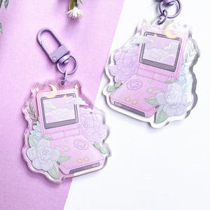 Puede incluir: Dos llaveros Game Boy Color rosa y morado con un diseño de luna y estrellas. Los llaveros están hechos de acrílico transparente y presentan un diseño floral.