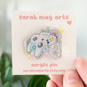 Puede incluir: Un mando de Nintendo 64 blanco con detalles florales, rodeado de flores de color rosa pastel, azul y verde. El texto "sarah may arts" y "acrylic pin" es visible en la tarjeta. La dirección del sitio web "sarahmayarts.etsy.com" también es visible.