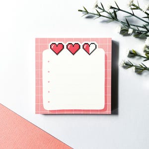 May include: Pink notepad with a grid pattern and three pixelated hearts at the top. The hearts are red and white. The notepad has a white space for writing.