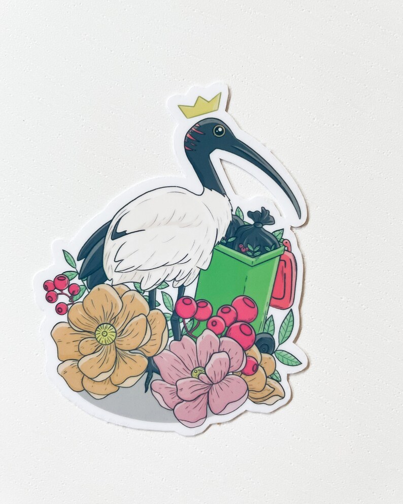 May include: A white ibis wearing a gold crown holds a green bin with a black bag inside. The ibis is surrounded by pink and yellow flowers and red berries.