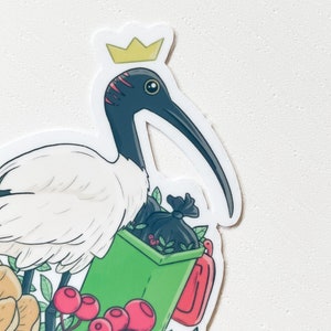 May include: A white ibis wearing a gold crown stands next to a green bin with a black bag full of leaves. The ibis is surrounded by pink, yellow, and green flowers.