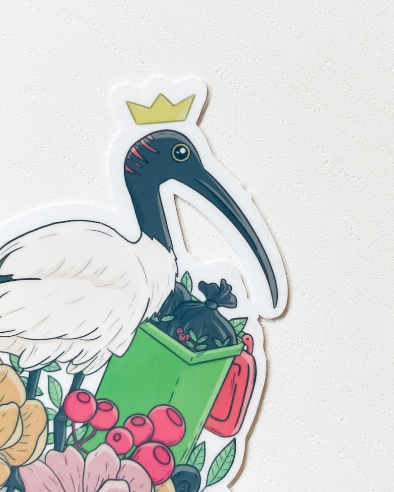 May include: A white ibis wearing a gold crown stands next to a green bin with a black bag full of leaves. The ibis is surrounded by pink, yellow, and green flowers.