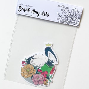 May include: A sticker featuring a white ibis wearing a gold crown, holding a green bin with black bags inside. The ibis is surrounded by pink, red, and yellow flowers.